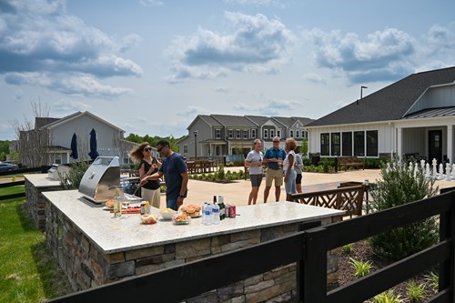 Active adults gathered around an outdoor grill and patio, enjoying food and drinks in a suburban community setting.