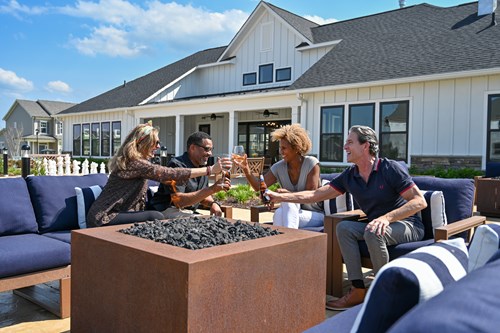 Residents gathered around an outdoor fire pit, toasting drinks in a community lounge area with clubhouse in the background.